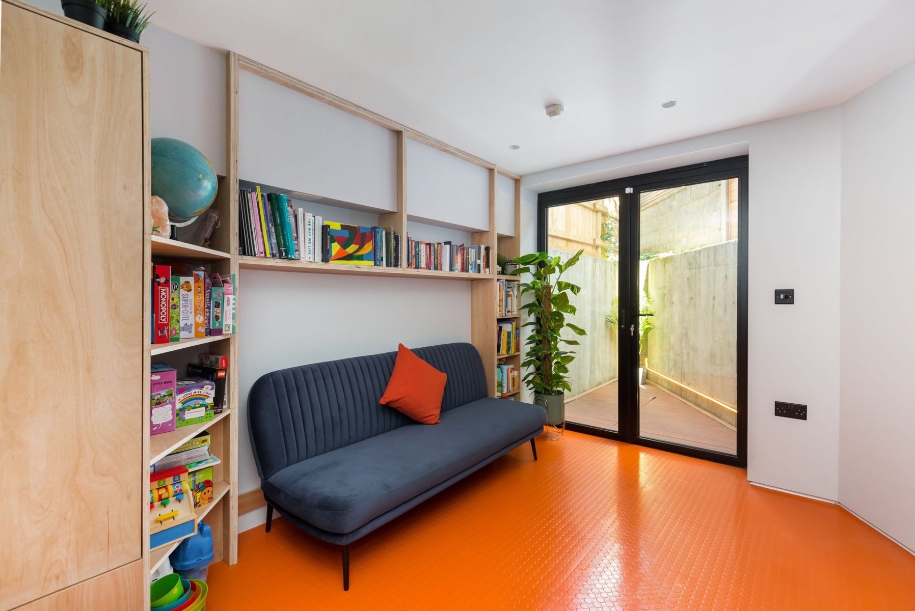 Living space with orange flooring, built-in timber shelving and glazed doors in a new-build house in Forest Hill.