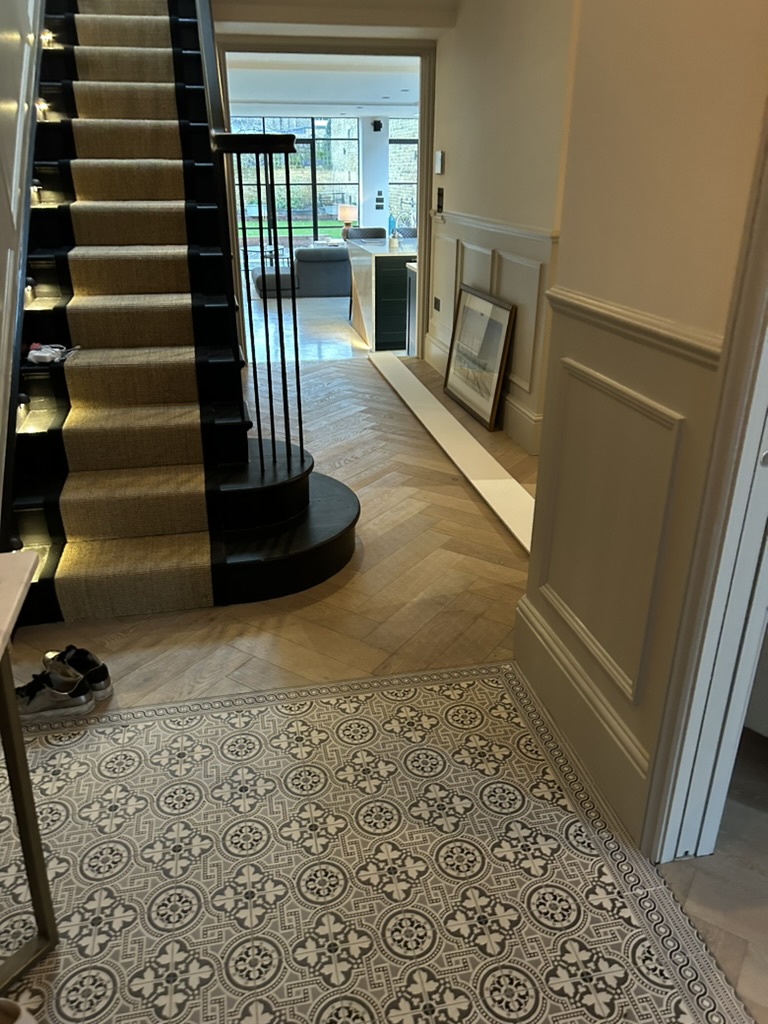 Widened Victorian hallway with curved staircase, black ironwork, herringbone oak floor meeting patterned tiles and wall panelling, Battersea SW11